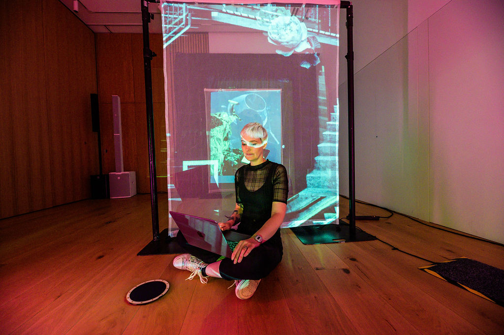 Photograph of me, a white woman in her 30s with a bleached pixie cut, sat in front of one of the projected collages in To Lie Within Another